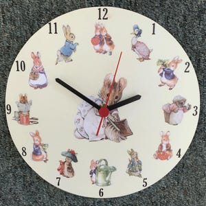 May include: A round wall clock with a cream-coloured face featuring black numerals from 1 to 12. The clock displays various illustrations of rabbits and a duck in clothing. A central image shows a rabbit sweeping with a broom. It has black hour and minute hands and a red second hand.