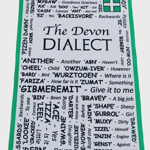 May include: A white rectangular tea towel with a green border featuring the text The Devon Dialect in large black letters. The towel lists various Devon dialect words and their meanings in black text, including Arken, Rufazrats, and Gibmeremit.