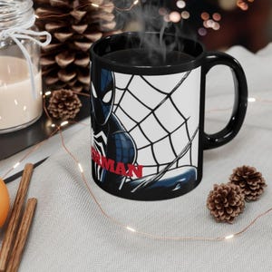 May include: Black ceramic mug featuring a Spider-Man graphic. The design includes a black and blue Spider-Man figure with a white spider web background and the word "SPIDERMAN" in red. The mug has a black interior and handle.
