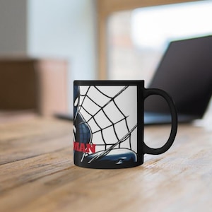 May include: Black ceramic mug featuring a comic book illustration of Spider-Man in a web. The word "MAN" is in red. The mug is on a wooden surface with a laptop in the background.