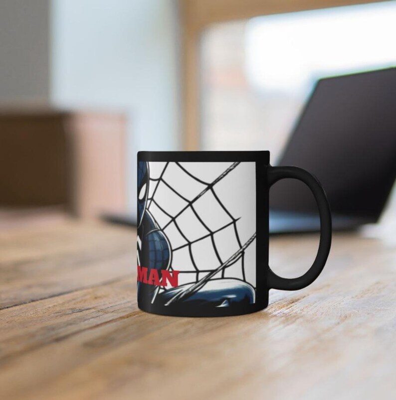 May include: Black ceramic mug featuring a comic book illustration of Spider-Man in a web. The word "MAN" is in red. The mug is on a wooden surface with a laptop in the background.
