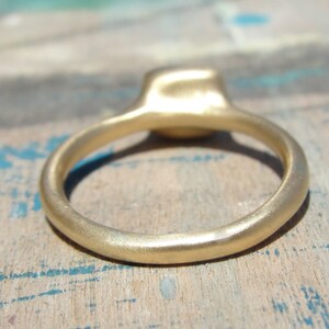 Op de afbeelding: Een gouden ring met een gladde, matte afwerking en een afgeronde band. De ring heeft een centrale zetting die iets boven de cirkelvormige band uitsteekt, wat een consistente metalen textuur over het hele sieraad laat zien.