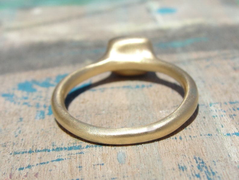Op de afbeelding: Een gouden ring met een gladde, matte afwerking en een afgeronde band. De ring heeft een centrale zetting die iets boven de cirkelvormige band uitsteekt, wat een consistente metalen textuur over het hele sieraad laat zien.