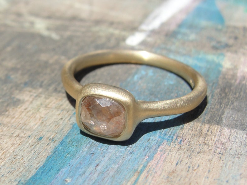Op de afbeelding: Een gouden ring met een matte afwerking, voorzien van een enkele, kussengeslepen, doorschijnende, lichtbruine edelsteen in een vierkante zetting. De ringband is glad en afgerond en vertoont een consistente metalen textuur over het geheel.