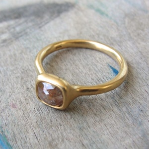Op de afbeelding: Een gouden ring met een gladde, mat afgewerkte band en een enkele, kussengeslepen, doorschijnende bruine edelsteen in een zetting. Het metaal heeft een warme, gele tint en de steen vertoont een gefacetteerd oppervlak met zichtbare insluitingen.
