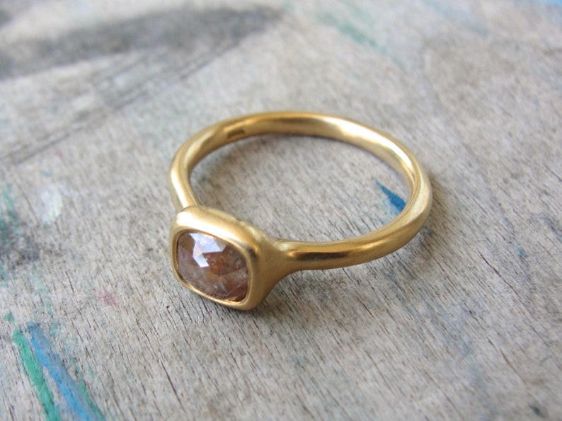 Op de afbeelding: Een gouden ring met een gladde, mat afgewerkte band en een enkele, kussengeslepen, doorschijnende bruine edelsteen in een zetting. Het metaal heeft een warme, gele tint en de steen vertoont een gefacetteerd oppervlak met zichtbare insluitingen.