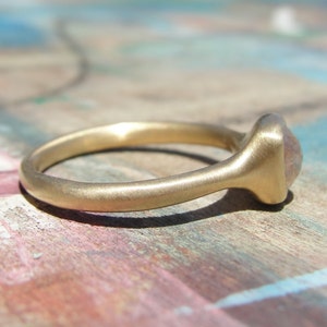 Op de afbeelding: Een gouden ring met een gladde, matte afwerking en een afgeronde band. De ring is voorzien van een enkele, kleine, doorschijnende, lichtgekleurde steen in een zetting aan de bovenzijde. Het metaal heeft een zachte, geborstelde textuur.