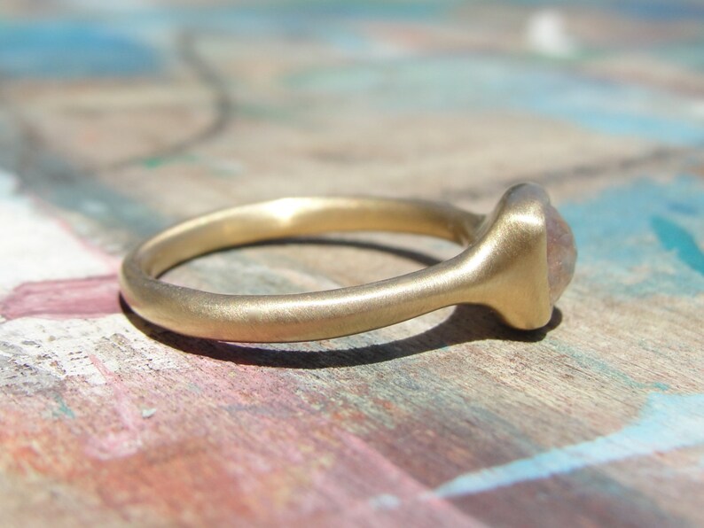 Op de afbeelding: Een gouden ring met een gladde, matte afwerking en een afgeronde band. De ring is voorzien van een enkele, kleine, doorschijnende, lichtgekleurde steen in een zetting aan de bovenzijde. Het metaal heeft een zachte, geborstelde textuur.