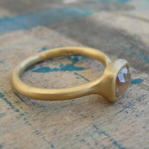 Op de afbeelding: Een gouden ring met een matte afwerking en een enkele, gefacetteerde, doorschijnende steen in een zetting. De ringband is afgerond en glad, met een zachte, niet-reflecterende textuur die contrasteert met de facetten van de centrale steen.