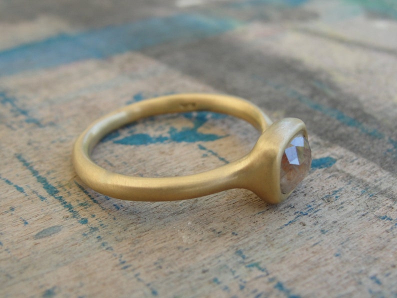 Op de afbeelding: Een gouden ring met een matte afwerking en een enkele, gefacetteerde, doorschijnende steen in een zetting. De ringband is afgerond en glad, met een zachte, niet-reflecterende textuur die contrasteert met de facetten van de centrale steen.