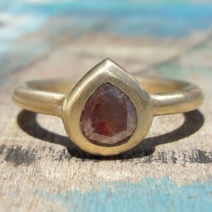 Op de afbeelding: Een gouden ring met een peervormige, roodbruine edelsteen in een gladde, mat afgewerkte gouden band. De steen heeft een gefacetteerd oppervlak en zit stevig in een zetting die de druppelvorm van de edelsteen nauwkeurig volgt.