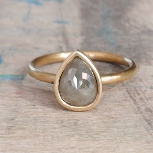 Op de afbeelding: Een gouden ring met een peervormige, gefacetteerde grijze steen in een gladde zetting. De ringband is dun en rond, gemaakt van gepolijst geelgoud. De steen heeft een doorschijnende, gedempte grijze kleur met zichtbare facetten in het oppervlak.