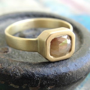 Op de afbeelding: Een gouden ring met een matte afwerking, voorzien van een vierkante, gefacetteerde bruine edelsteen in een gladde zetting. De ringband is glad en heeft een gelijkmatige breedte, wat zorgt voor een strak ontwerp dat het contrast benadrukt.