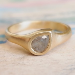 Op de afbeelding: Een gouden ring met een gladde, mat afgewerkte band en een enkele, peervormige, gefacetteerde grijze steen in een zetting. De ring heeft een stevige, afgeronde schacht die iets taps toeloopt naar de centrale steen toe.