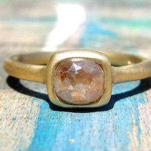 Op de afbeelding: Een gouden ring met een kussengeslepen, doorschijnende, lichtbruine edelsteen gezet in een gladde zetting met een matte afwerking. De ringband is van massief goud met een afgerond profiel en een zachte, niet-reflecterende textuur.