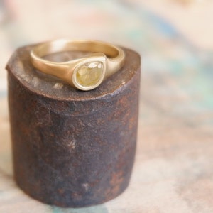 Op de afbeelding: Een gouden ring met een gladde, mat afgewerkte band en een enkele, in een zetting geplaatste, peervormige gele diamant. De steen heeft een gefacetteerd oppervlak en zit stevig vast in de metalen zetting die naadloos overloopt in de band.