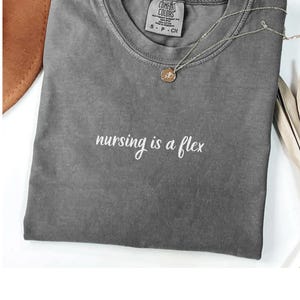 May include: A gray Comfort Colors t-shirt with the phrase "nursing is a flex" in white script. A silver necklace with a small pendant rests on the shirt. The shirt is folded and placed on a white surface.