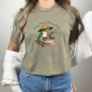 May include: Olive green t-shirt featuring a cartoon frog wearing a conical hat and eating noodles from a bowl with chopsticks. The text "Slurp First, Worry Later" is arched above the frog. The shirt is made of a soft material.