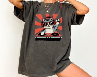 May include: Dark gray t-shirt featuring a cartoon cat DJ wearing sunglasses and headphones, waving from behind a turntable. The cat is set against a red sunburst design. The shirt has short sleeves and a relaxed fit.