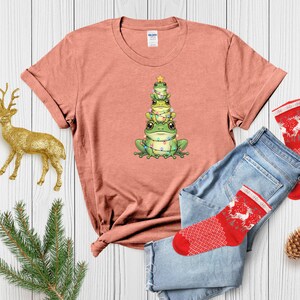 May include: A coral-colored t-shirt featuring a graphic of three green frogs stacked like a Christmas tree, adorned with string lights and a star. The shirt is paired with blue jeans and red patterned socks, suggesting a festive theme.