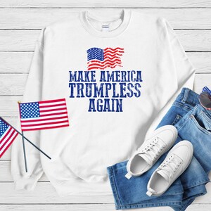 May include: White sweatshirt with the text "MAKE AMERICA TRUMPLESS AGAIN" in blue, along with a distressed American flag graphic. The image also includes a pair of blue jeans, white sneakers, and small American flags.