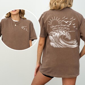 May include: A brown short-sleeve t-shirt featuring a white graphic on the back of a large ocean wave, a sun, and birds with the text "When you go through deep waters, i will be with you Isaiah 43:2". The front chest has a small bird graphic.