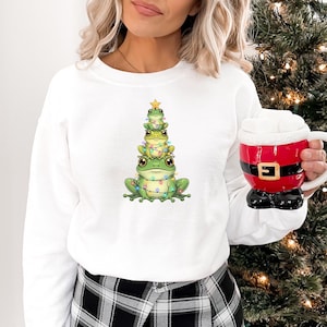 May include: White sweatshirt featuring a whimsical design of three green frogs stacked like a Christmas tree, adorned with string lights and a star. The frogs are the focal point, creating a festive and playful aesthetic. A Santa-themed mug is held in the hand.