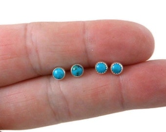 May include: Three pairs of small, round turquoise stud earrings with silver settings. The earrings are shown on a finger.