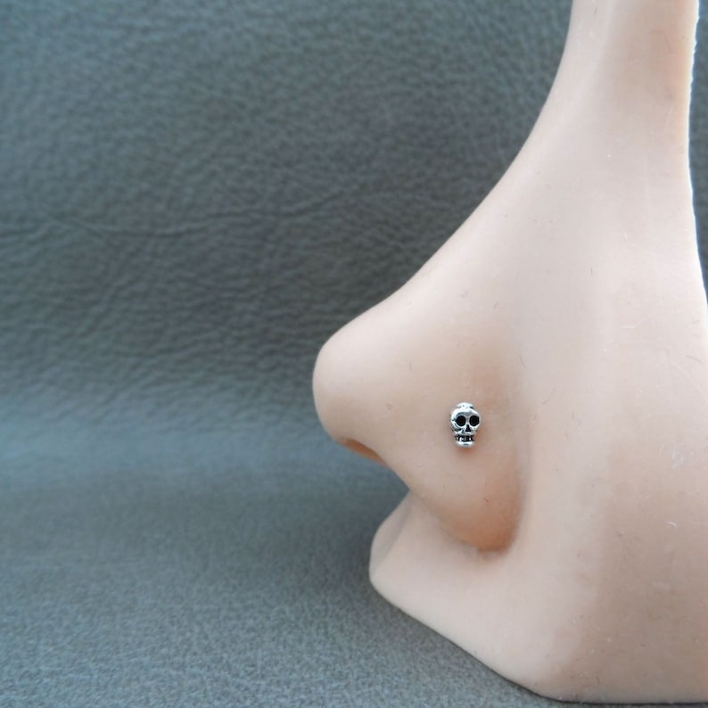 K&ouml;nnte beinhalten: Ein kleiner Nasenpiercing-Stecker in Sch&auml;delform, silberfarben. Der Sch&auml;del hat schwarze Details f&uuml;r Augen und Nase. Der Stecker wird auf einer Schaufensternase pr&auml;sentiert, um Gr&ouml;&szlig;e und Design zu zeigen.