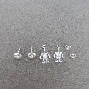 May include: A set of six silver earrings featuring two skull designs and two skeleton designs. The earrings are all silver and have a shiny finish.