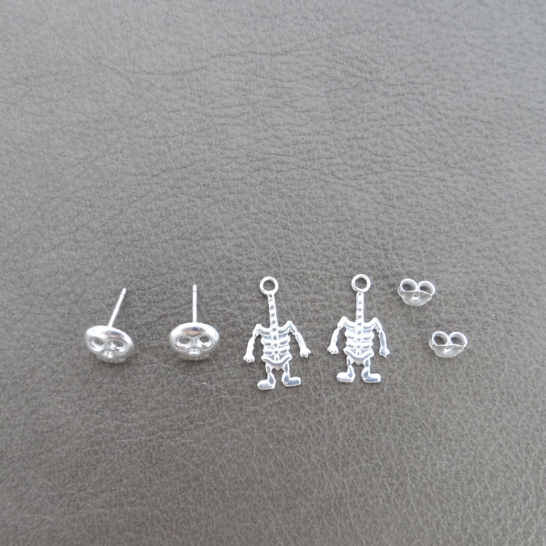 May include: A set of six silver earrings featuring two skull designs and two skeleton designs. The earrings are all silver and have a shiny finish.