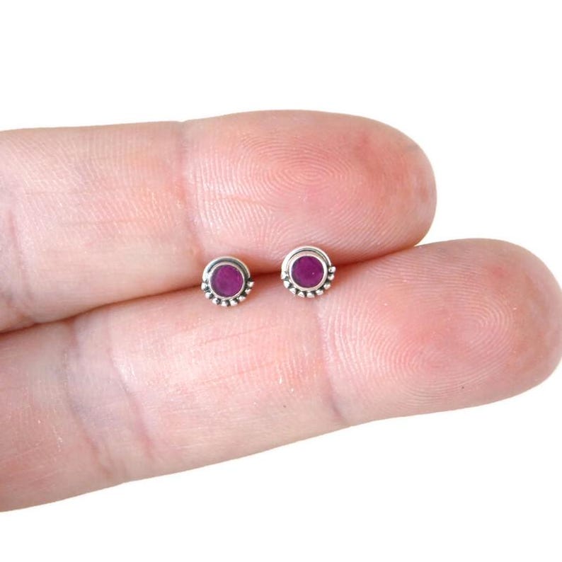 May include: A pair of small, silver stud earrings with a round, purple gemstone in the centre of each earring.