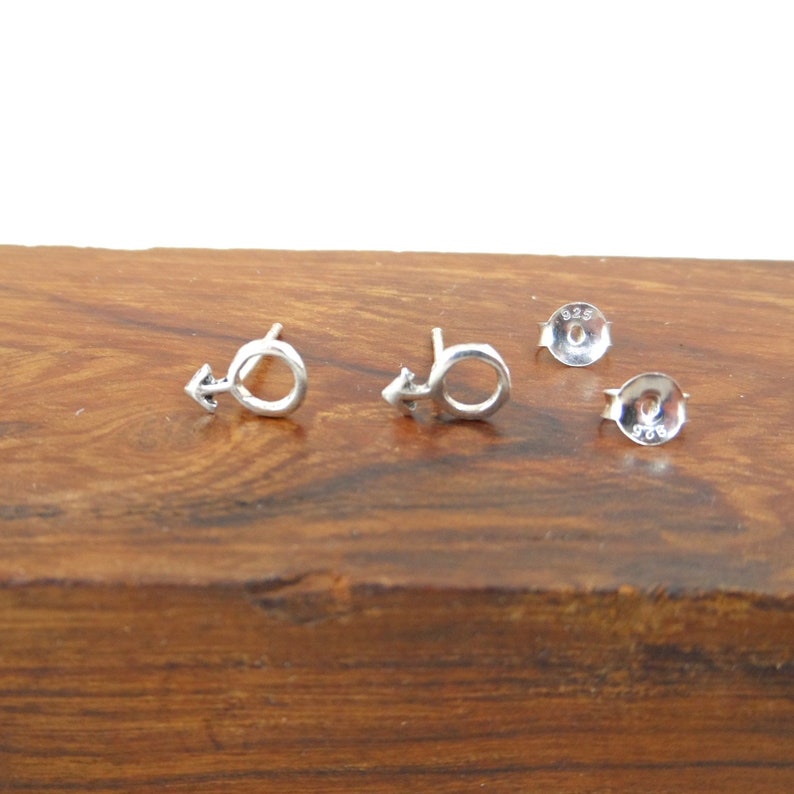 May include: A pair of silver earrings with a male symbol design. The earrings are on a wooden surface.