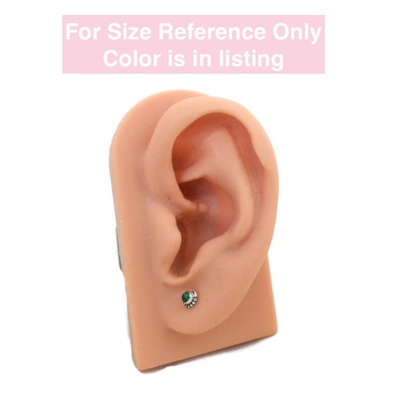May include: A close-up of a fake ear with a small green gemstone earring. The earring is silver with a decorative design around the stone.