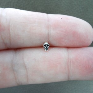 K&ouml;nnte beinhalten: Ein kleiner, silberfarbener Totenkopf-Anh&auml;nger mit detaillierten Augenh&ouml;hlen und Kiefer. Der Anh&auml;nger wird zwischen zwei Fingern gehalten, was seine Miniaturgr&ouml;&szlig;e und sein aufwendiges Design zeigt. Dieser Anh&auml;nger eignet sich f&uuml;r die Schmuckherstellung.