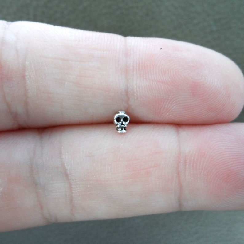 K&ouml;nnte beinhalten: Ein kleiner, silberfarbener Totenkopf-Anh&auml;nger mit detaillierten Augenh&ouml;hlen und Kiefer. Der Anh&auml;nger wird zwischen zwei Fingern gehalten, was seine Miniaturgr&ouml;&szlig;e und sein aufwendiges Design zeigt. Dieser Anh&auml;nger eignet sich f&uuml;r die Schmuckherstellung.