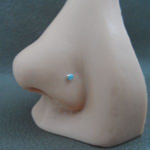 May include: A close-up of a light blue gemstone nose ring stud. The stud is set in a silver setting and is worn on a model's nose.