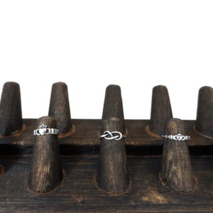 May include: A wooden ring display with three silver rings. The rings are on individual wooden posts. The rings are a Celtic knot, a Claddagh ring, and a heart ring.