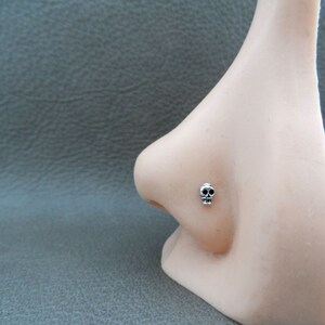 K&ouml;nnte beinhalten: Ein silberfarbener Nasenpiercing mit Totenkopf. Der kleine Totenkopf hat schwarze Augenh&ouml;hlen und ein detailliertes Design. Der Nasenpiercing wird an einer Schaufensternase gezeigt.