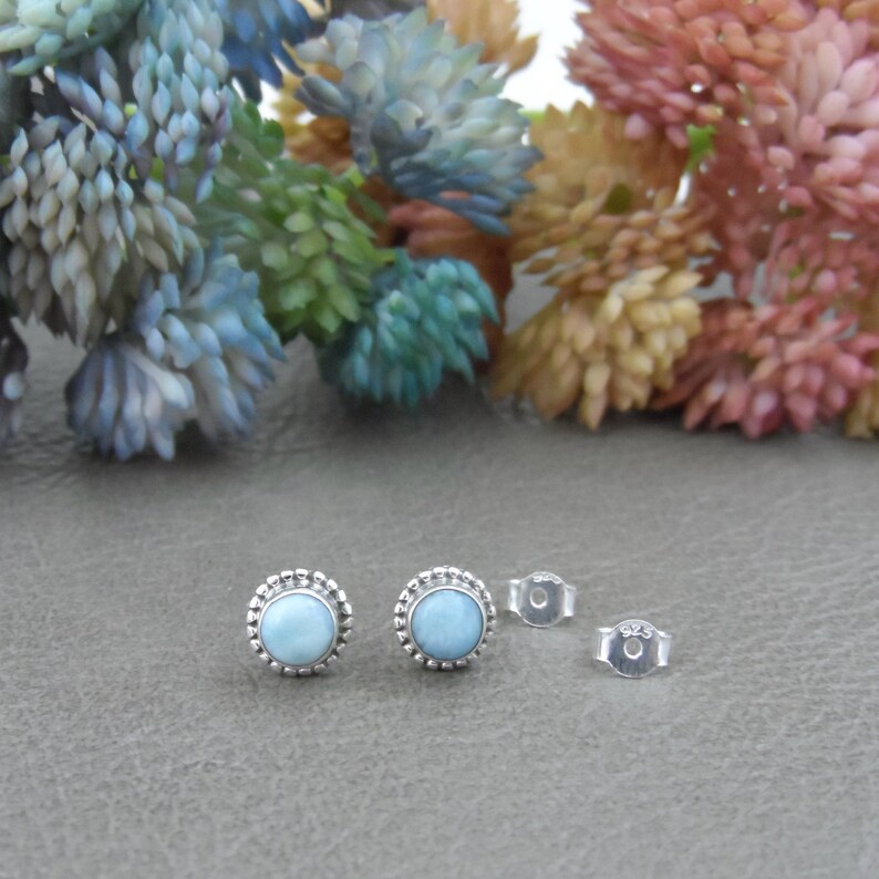 May include: A pair of silver earrings with light blue gemstone cabochons set in a beaded bezel. The earrings have a simple, elegant design.
