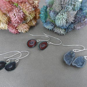 May include: Three pairs of silver dangle earrings with different colored agate slices. The earrings feature a silver hook and a round agate slice with a natural geode pattern. The agate slices are red, green, and blue.