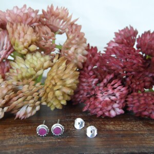 May include: A pair of silver stud earrings with small pink gemstones set in the centre. The earrings are displayed on a wooden surface with pink flowers.
