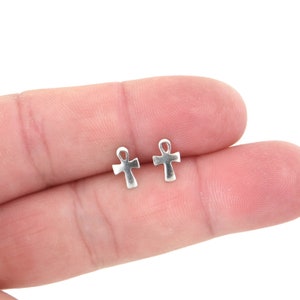 Könnte beinhalten: Zwei kleine, silberfarbene Ohrringe in Ankh-Form. Das Ankh ist ein altägyptisches Hieroglyphensymbol, das das Leben darstellt. Die Ohrringe werden auf dem Finger einer Person gezeigt.