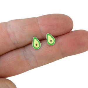May include: A pair of avocado-shaped stud earrings. Each earring features a green enamel outline, a yellow-green center, and a small brown seed. The earrings have a silver-colored metal border.