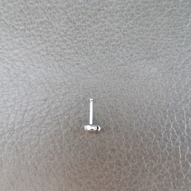 K&ouml;nnte beinhalten: Ein kleiner, silberfarbener Ohrstecker mit Stecker und Zierelement. Der Ohrring ist vor einem neutralen grauen Hintergrund platziert.