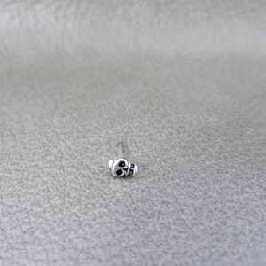K&ouml;nnte beinhalten: Ein kleiner, silberfarbener Nasenpiercing mit einem Totenkopf-Design. Der Totenkopf hat schwarze Augenh&ouml;hlen und einen detaillierten Kiefer. Der Stecker befindet sich auf einem d&uuml;nnen, geraden Stab.