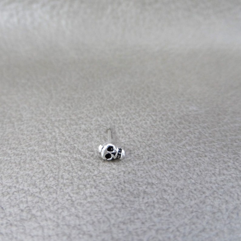 K&ouml;nnte beinhalten: Ein kleiner, silberfarbener Nasenpiercing mit einem Totenkopf-Design. Der Totenkopf hat schwarze Augenh&ouml;hlen und einen detaillierten Kiefer. Der Stecker befindet sich auf einem d&uuml;nnen, geraden Stab.