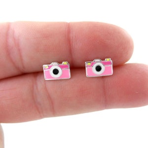 Puede incluir: Un par de pendientes de botón de cámara rosa con lente negra y detalles amarillos.