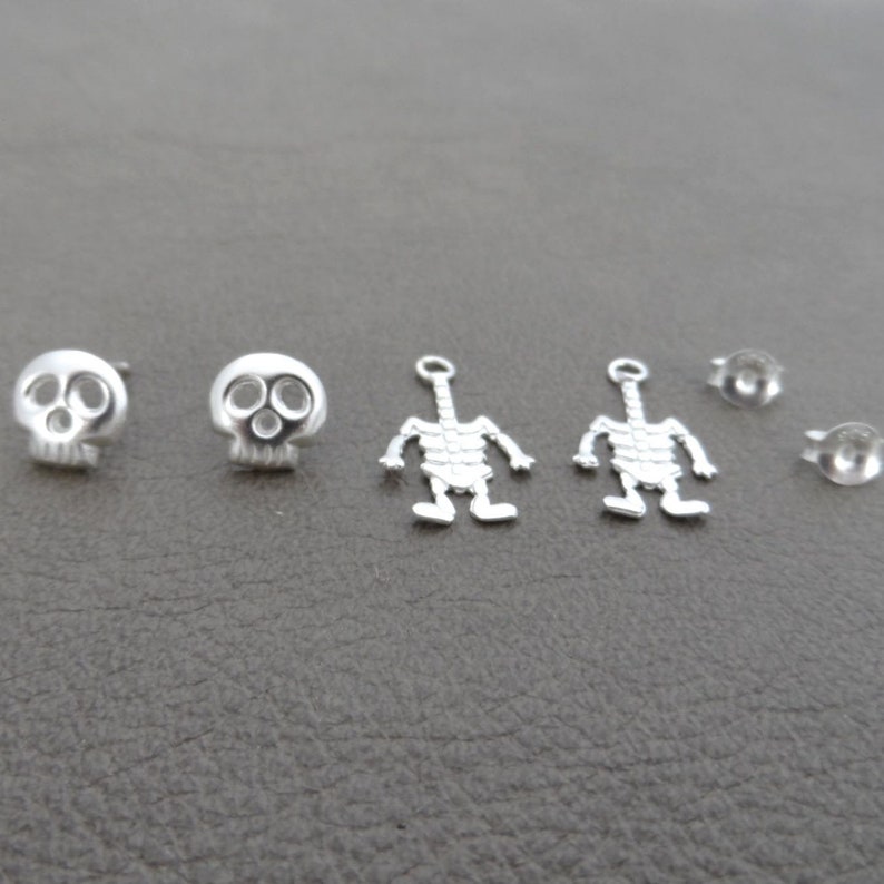 May include: A set of six silver earrings featuring two skull designs and two skeleton designs. The set also includes two earring backs.