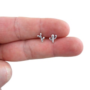 May include: A pair of silver-colored cactus stud earrings. One earring features a cluster of rounded shapes, while the other has a more traditional cactus silhouette. The earrings are small and delicate, perfect for everyday wear.